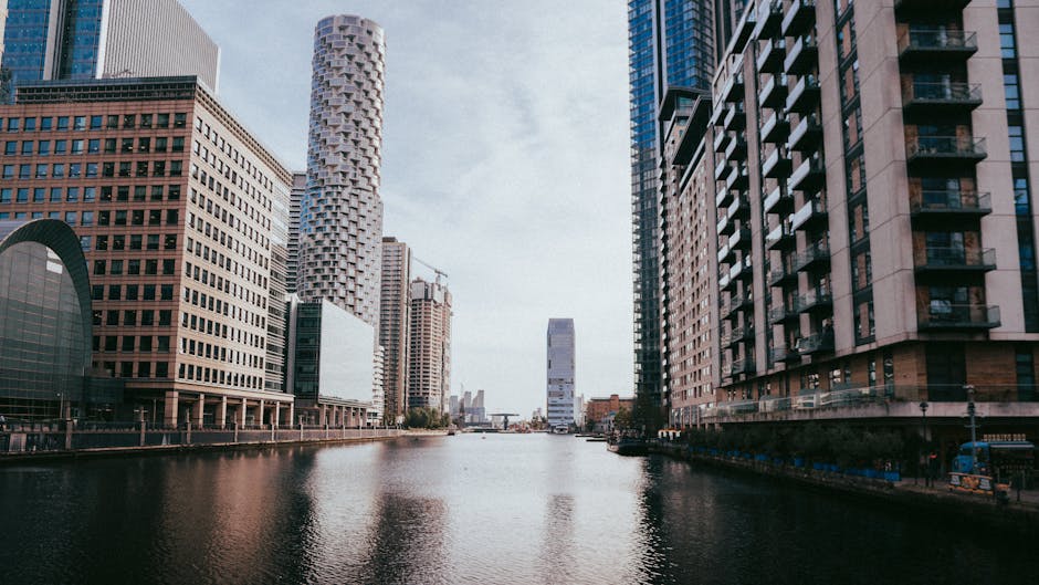 A wide view of the Canary Wharf skyline in daylight, showing modern high-rise buildings with glass and steel facades along a waterway. The buildings feature reflective glass windows, some with balconies, and are arranged beside a calm river surface with gentle ripples. The sky is partly cloudy, providing natural lighting that highlights the urban architecture. No cleaning tools or surfaces are visible, as this is an exterior cityscape image. Canary Wharf Cleaner specializes in surface and deep cleaning, ensuring hygiene and maintenance for residential and commercial premises in the area.