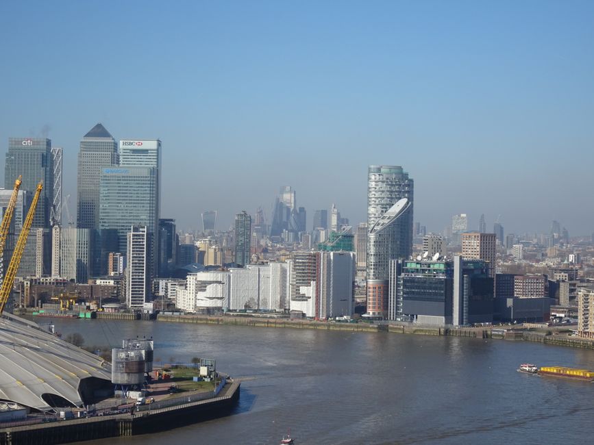 A wide view of the Canary Wharf skyline in daylight, showing modern high-rise buildings with glass and steel facades along a waterway. The buildings feature reflective glass windows, some with balconies, and are arranged beside a calm river surface with gentle ripples. The sky is partly cloudy, providing natural lighting that highlights the urban architecture. No cleaning tools or surfaces are visible, as this is an exterior cityscape image. Canary Wharf Cleaner specializes in surface and deep cleaning, ensuring hygiene and maintenance for residential and commercial premises in the area.