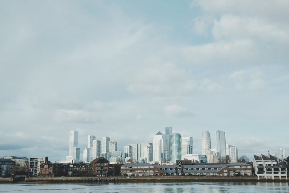 A wide view of the Canary Wharf skyline in the Docklands area during the late afternoon, featuring tall modern skyscrapers with glass facades reflecting the sunlight. Several construction cranes are p