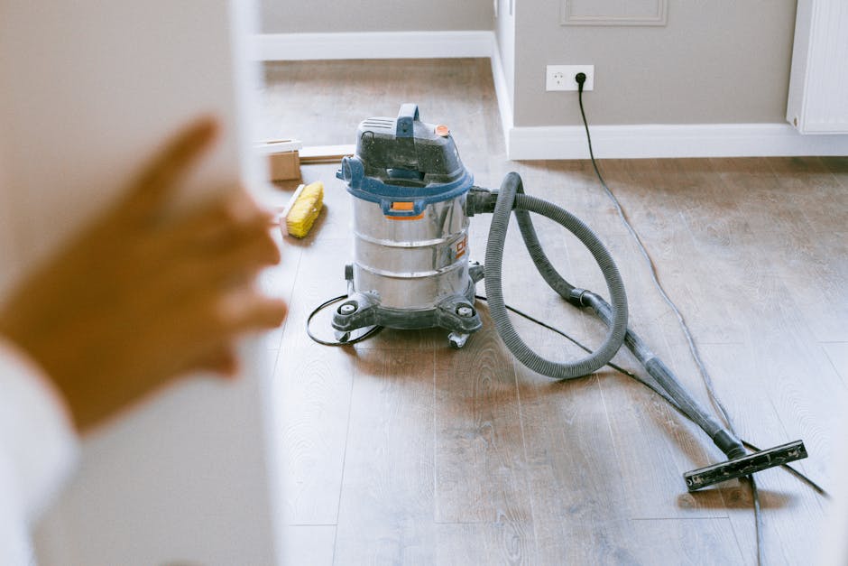 A room undergoing surface cleaning with a deep cleaning machine positioned on a wooden floor near a green-painted wall. The machine has a yellow body with a grey hose, and cleaning tools are visible. 