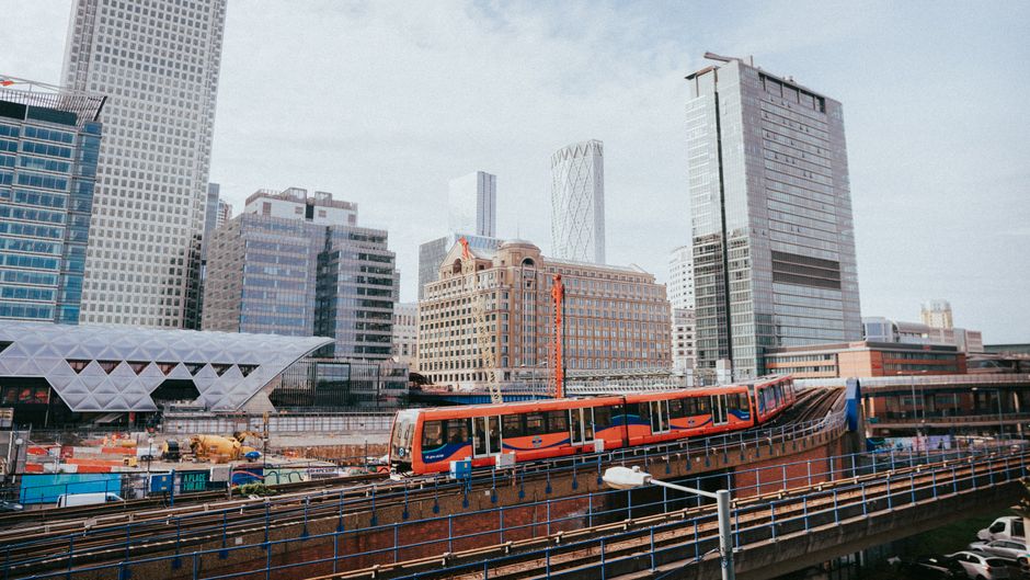 A high-rise office building with a modern glass façade featuring large rectangular windows and metal framing, situated in Canary Wharf. Two window cleaning technicians are on a suspended platform cle